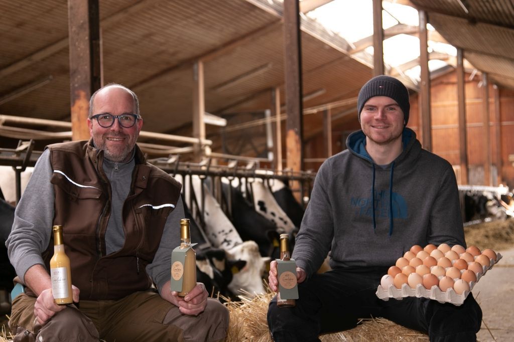 Zwei Männer in einer Scheune, die Flaschen und ein Tablett mit Eiern halten; im Hintergrund sind Kühe zu sehen.