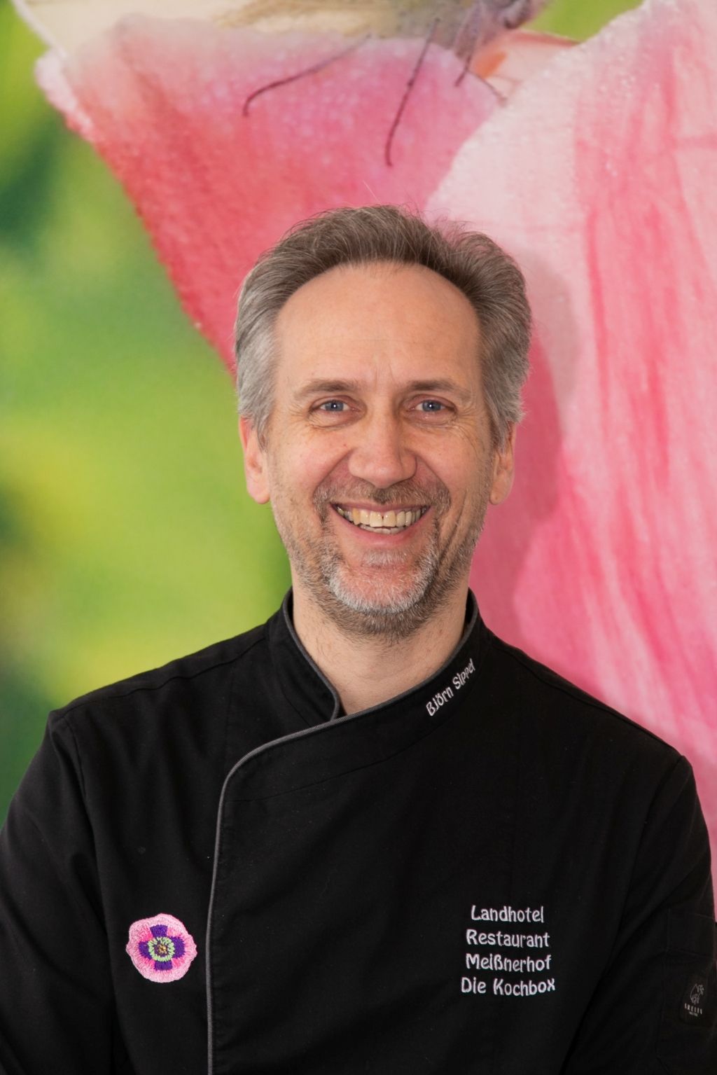 Der Koch im schwarzen Kochmantel lächelt. Das Logo des Landhotel-Restaurants „Waldkater“ ist auf dem Mantel zu sehen. Rosa-grüner Hintergrund.