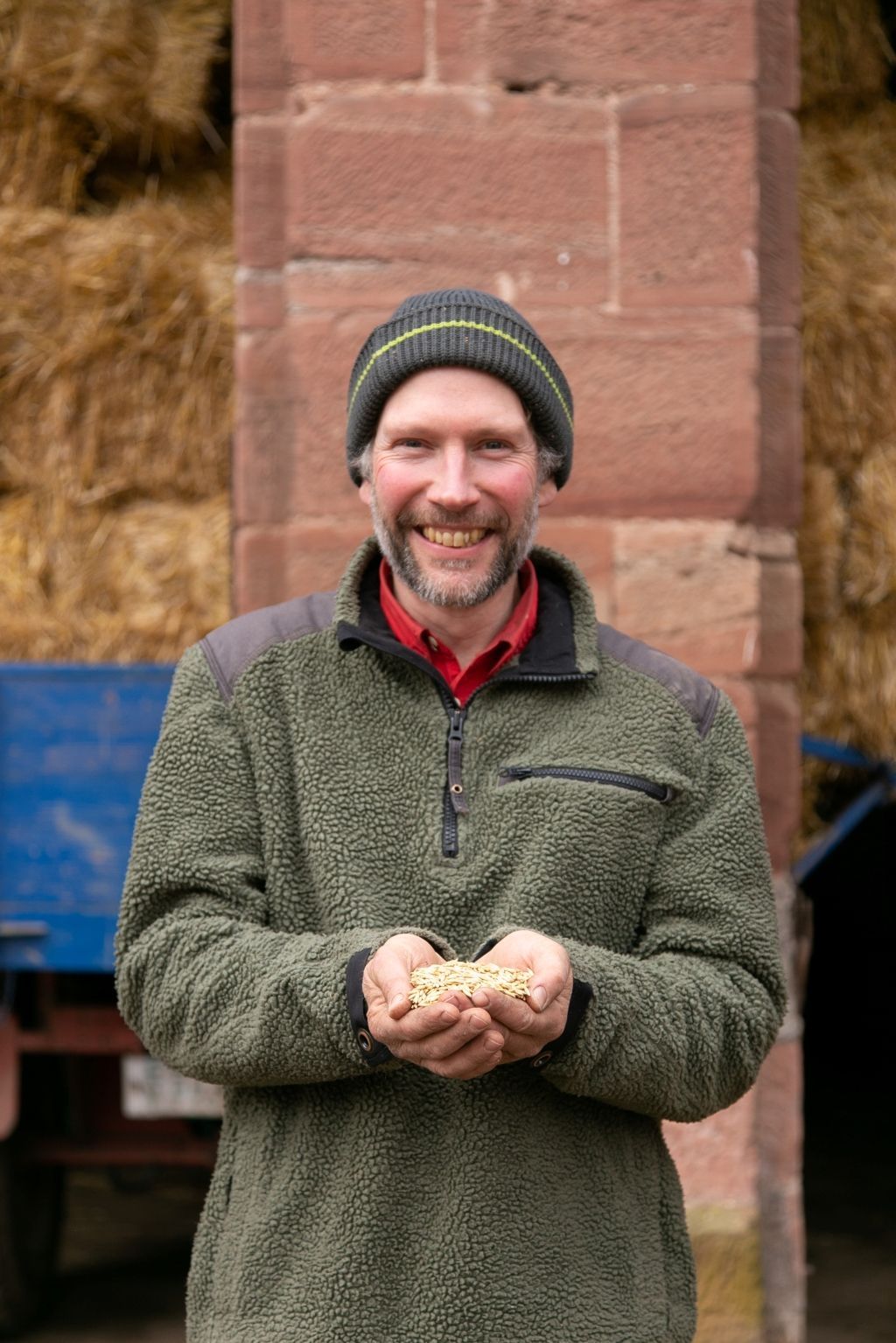 Ein Mann lächelt und hält Körner in der Hand. Er trägt eine Mütze und eine Fleecejacke und steht neben einem Heuballen und einem roten Ziegelstein.