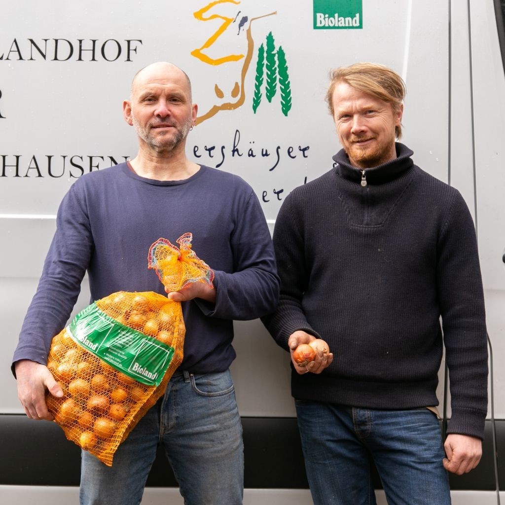 Zwei Männer mit Taschen und losen Zwiebeln stehen vor einem Lieferwagen mit Logo.