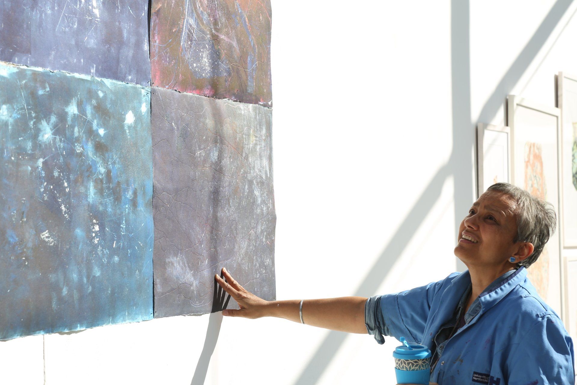Woman examining a square artwork of colored, textured panels on a white wall.