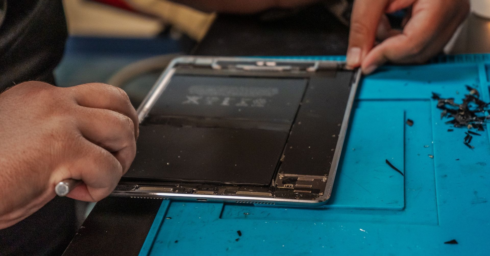 A person repairing a tablet, using tools to remove the screen from the frame on a blue mat.