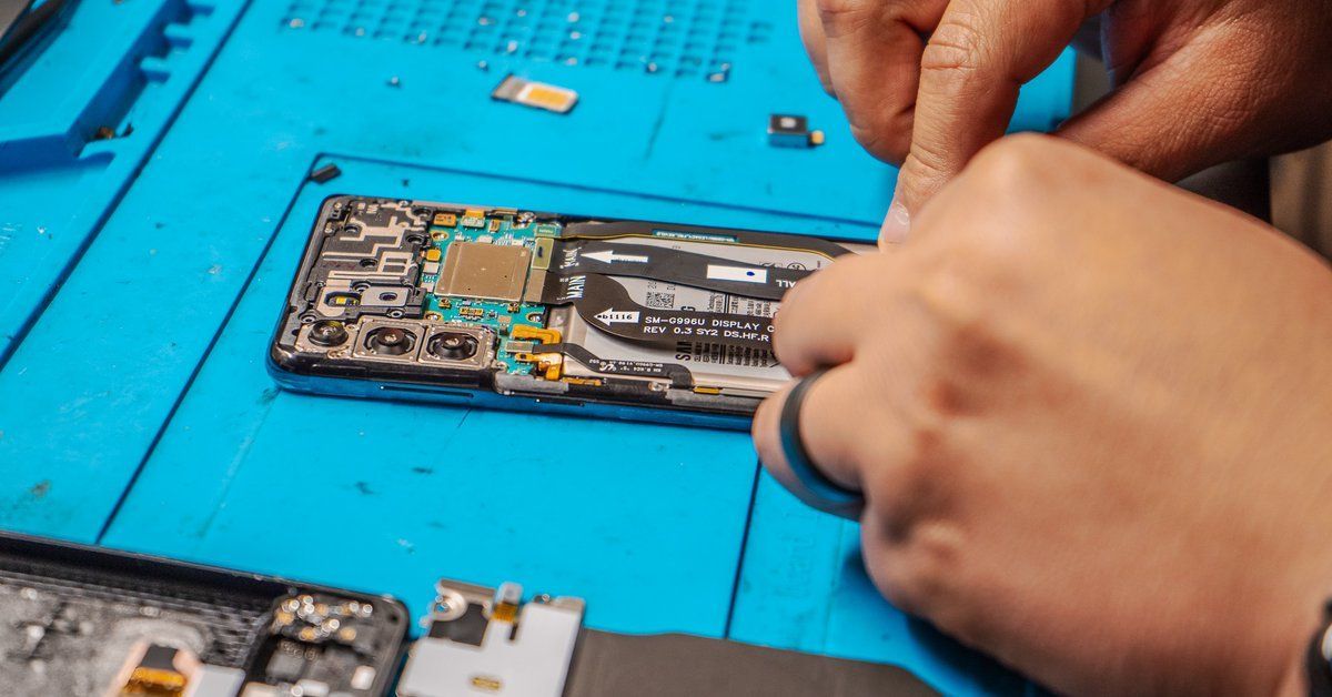 Hands working on the disassembled interior of a cell phone on a blue mat.