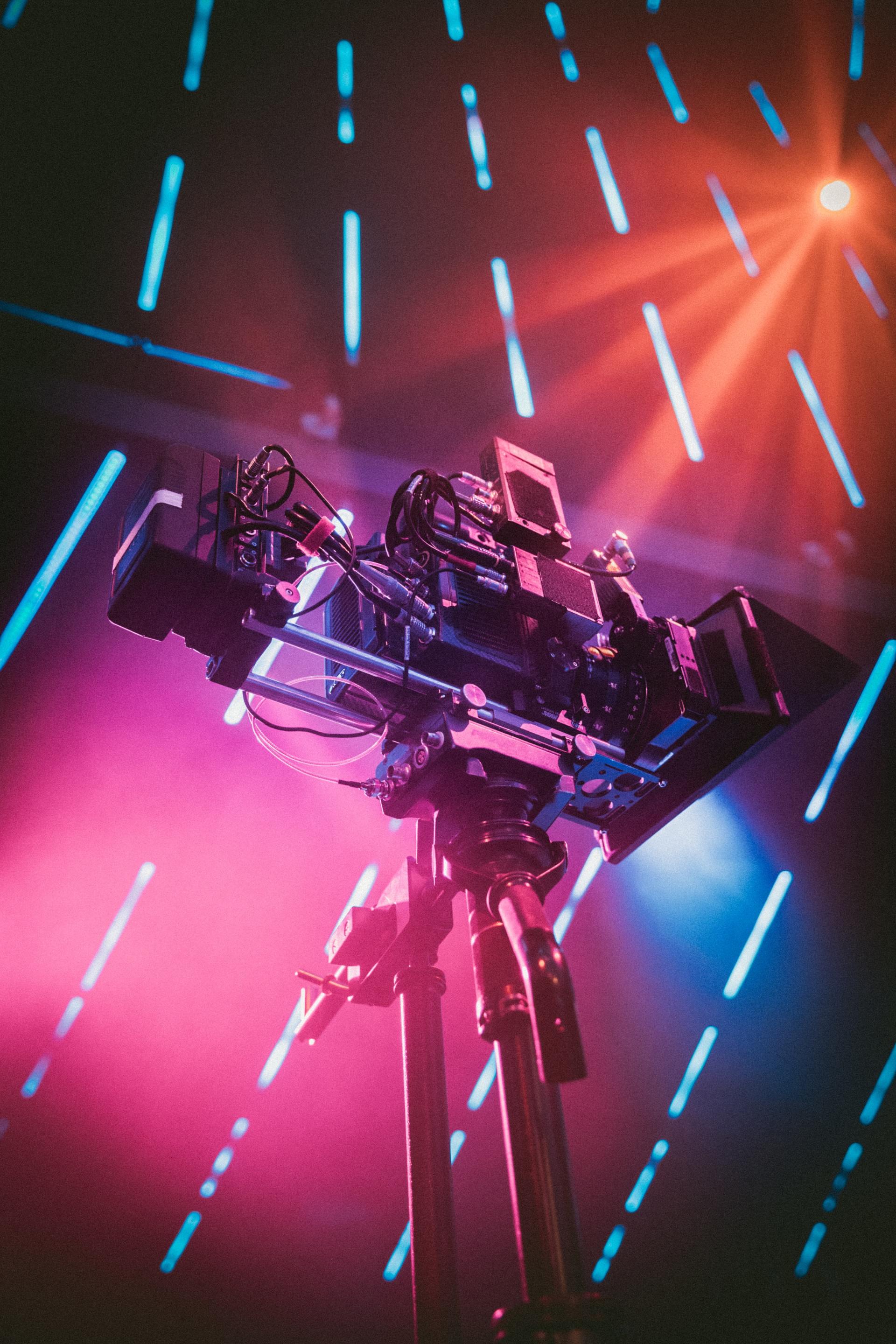 A complex camera rig mounted on a tripod, bathed in pink and blue stage lighting, with vertical light streaks.