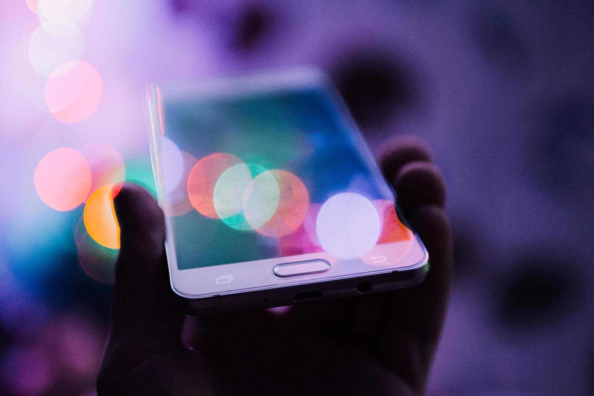 A hand holding a smartphone with a blurred background of colorful, out-of-focus lights.