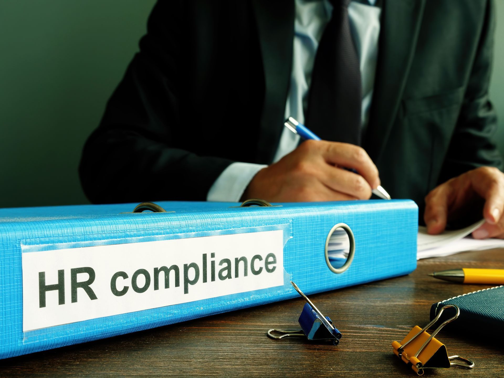 A man is sitting at a desk with a binder labeled hr compliance.