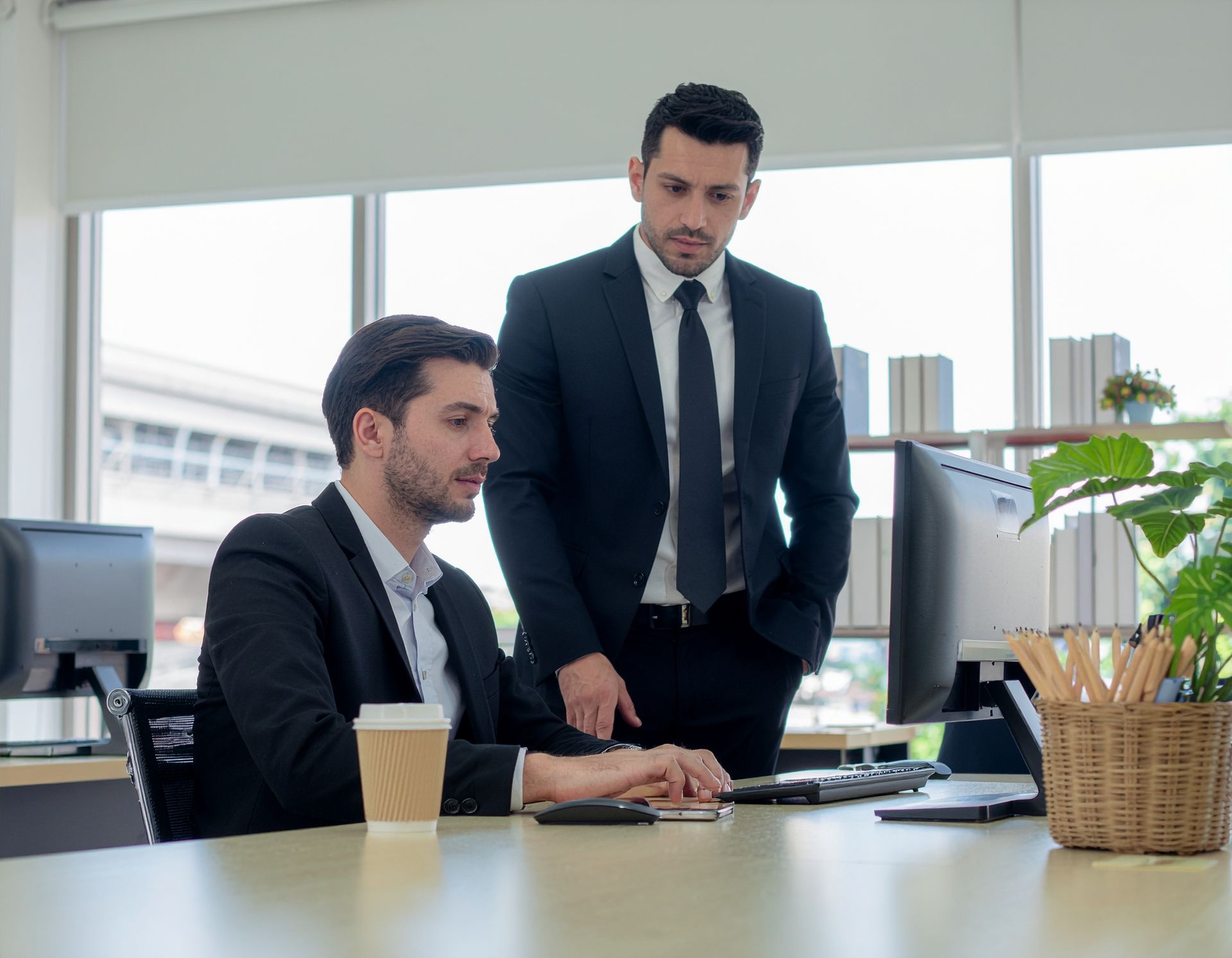 Two men in suits at a computer; one seated, the other standing and looking at the screen in a bright office.
