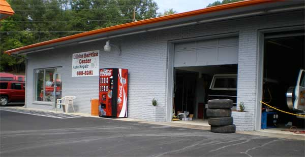 Exterior view of a gray auto service shop with an open garage bay and a Coca-Cola vending machine.