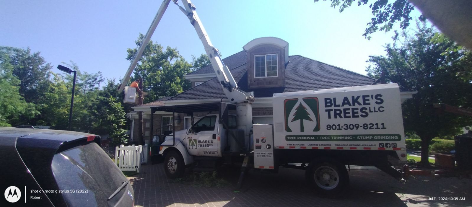 A tree service truck with a raised arm cutting a tree near a house. Blue sky.