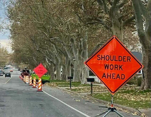 Orange road signs warn of shoulder work ahead, along a tree-lined street with traffic cones.