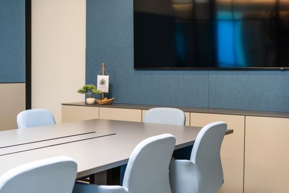 Konferenzraum mit grauem Tisch, hellblauen Stühlen und blauer Wand mit Fernseher.