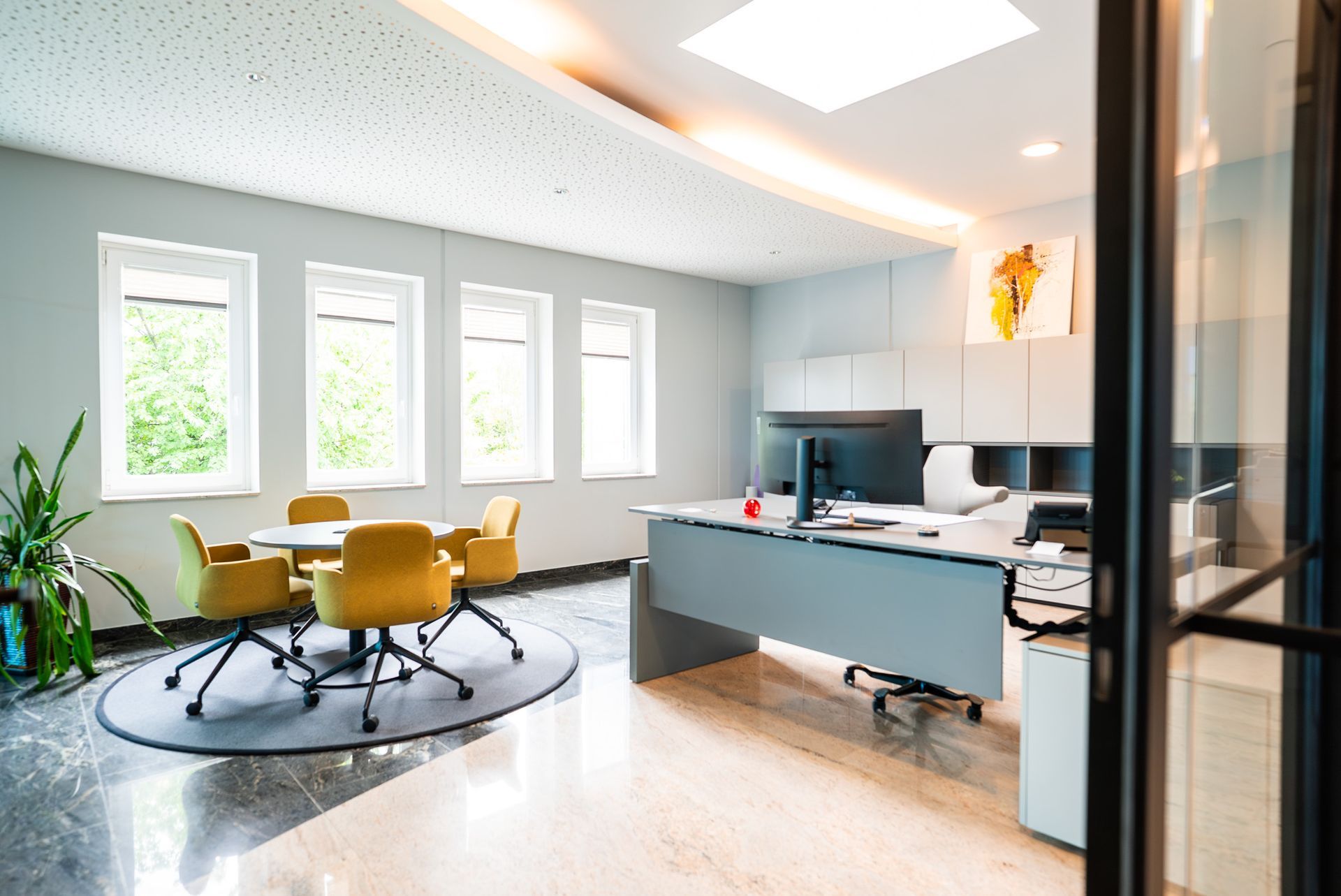 Büroeinrichtung mit Schreibtisch, rundem Tisch mit gelben Stühlen, Fenstern und Pflanzen.