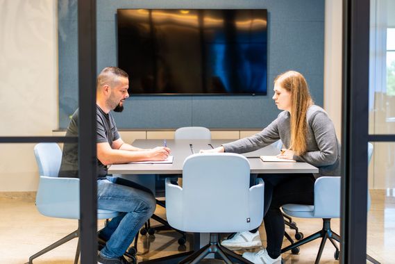 Zwei Personen sitzen in einem modernen Büro an einem Tisch und arbeiten zusammen. Die eine schreibt, die andere gestikuliert.