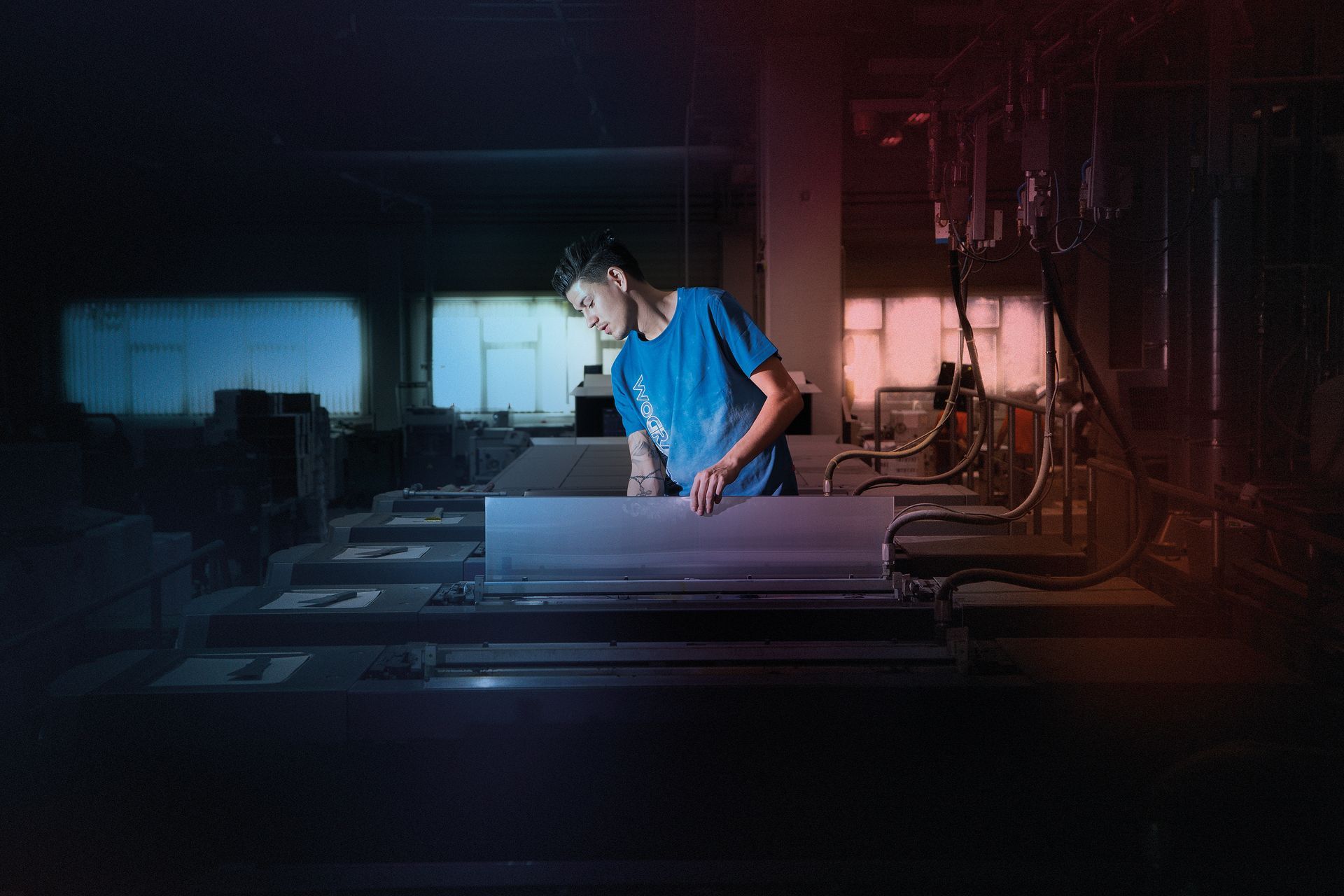 Person untersucht Metallgegenstand in einer schwach beleuchteten Industrieumgebung. Blaues Hemd, rot-blaue Beleuchtung.