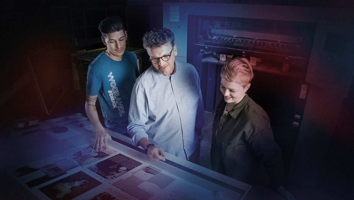 Drei Personen untersuchen gedruckte Materialien in einer Druckereiumgebung mit dunkelblauer und roter Beleuchtung.
