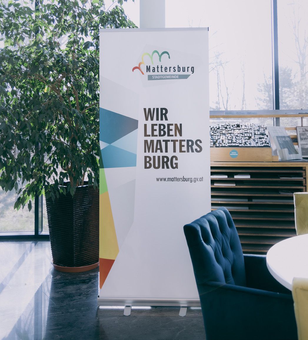 Banner: „Wir leben Mattersburg“ neben einer Topfpflanze und einem blauen Stuhl.