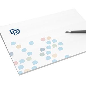 Weißer Notizblock mit blauem Logo und buntem Tropfenmuster, Stift an der Seite.