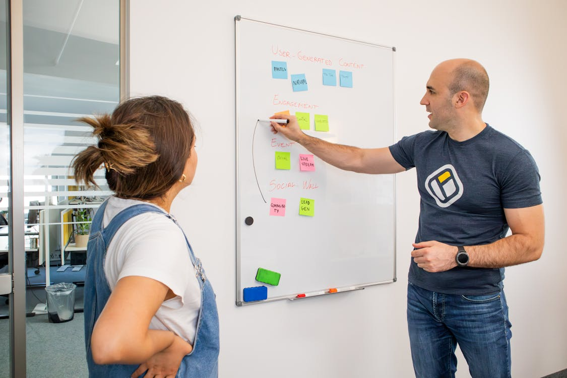 Two colleagues discussing priorities at a whiteboard, with one pointing to notes outlining tasks, engagement, and planning categories.
