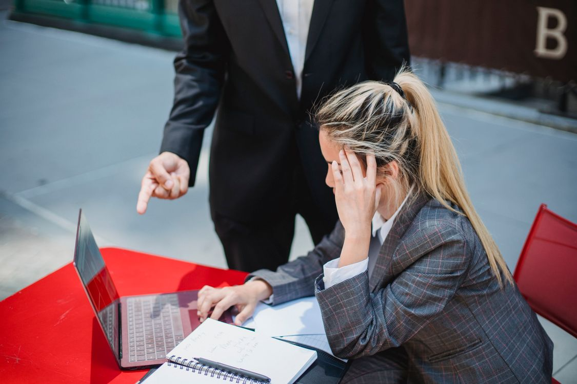 A stressed employee sitting at a desk working on a laptop while a manager points at her, highlighting workplace pressure, guidance, or performance correction in a professional office setting.
