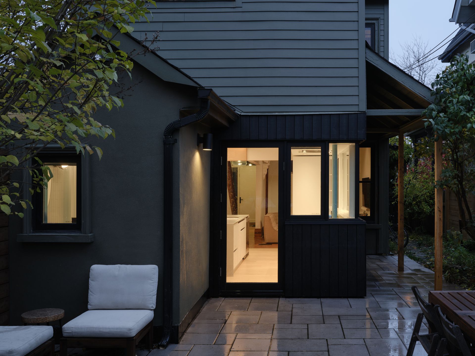 Dark-painted home exterior with an illuminated interior seen from a paved patio. Two white chairs and a table are on the patio.