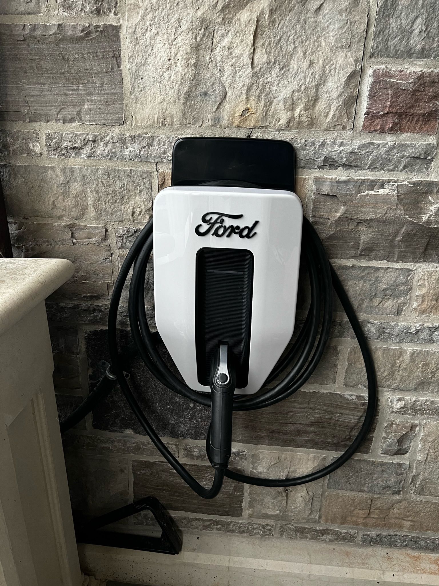 Ford EV charger mounted on a stone wall. Black charging cable coiled below the white charger.