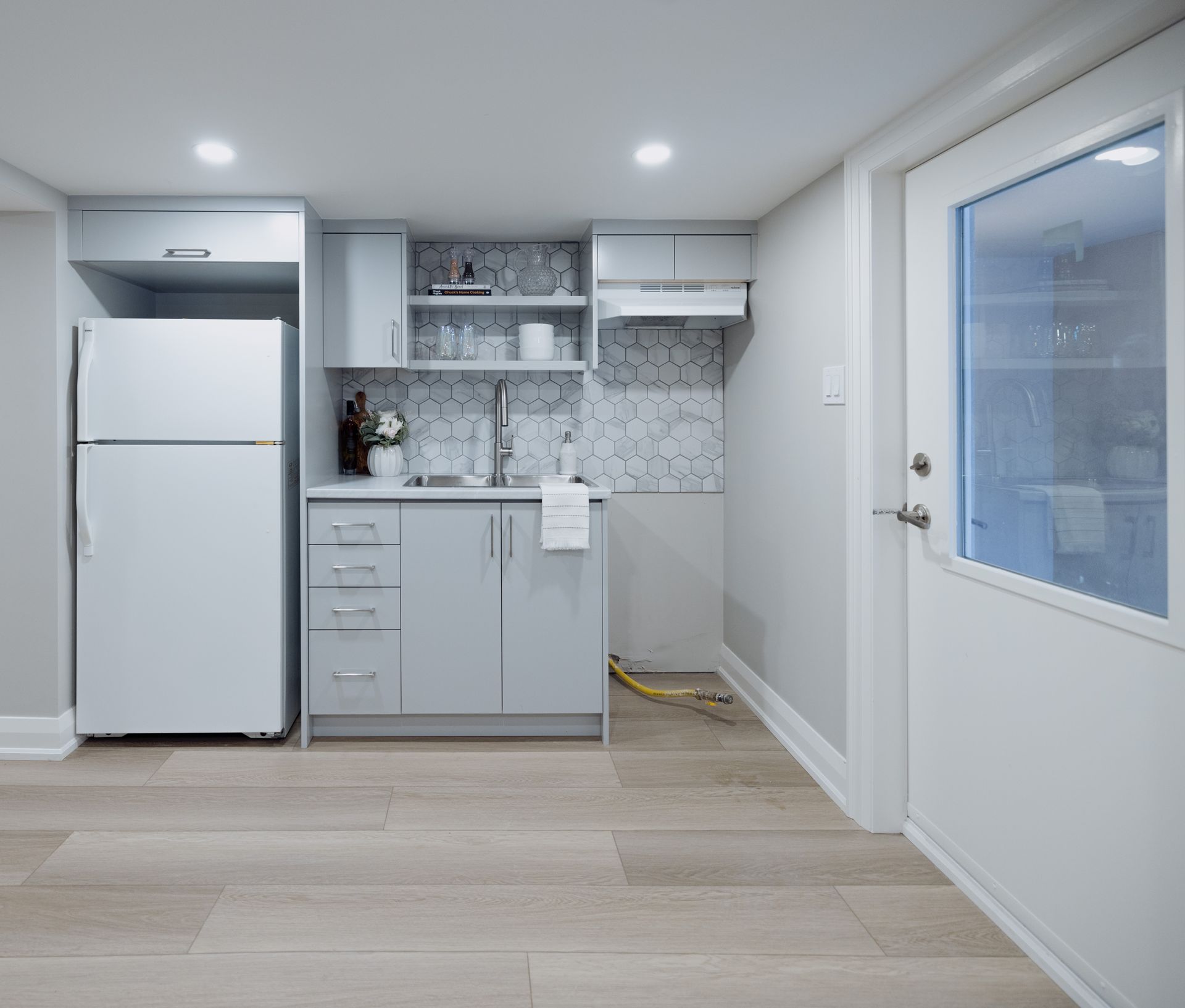 Small basement kitchen with light gray cabinets, white refrigerator, and a white door to the right.