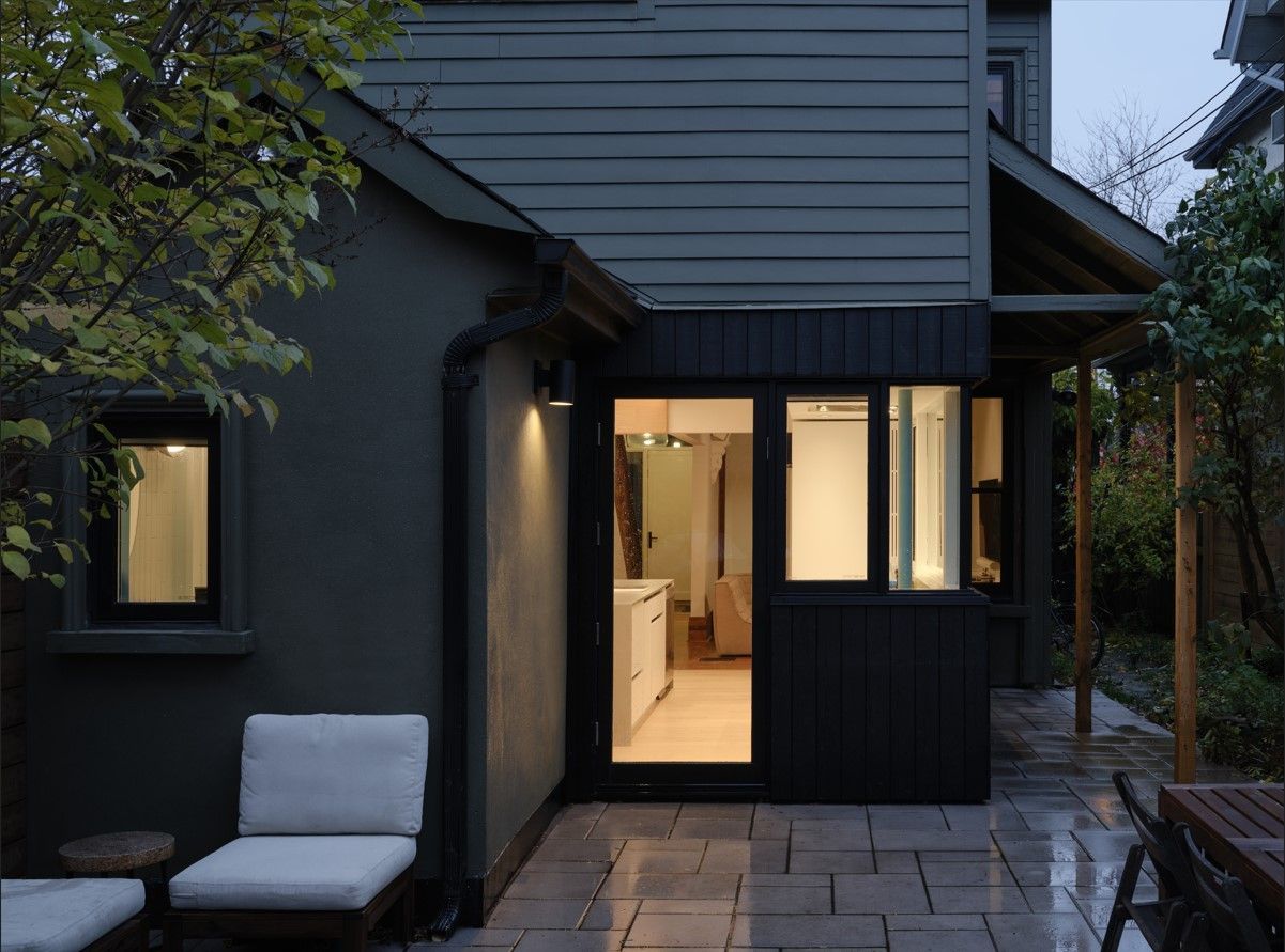 Dark gray house with illuminated windows and open doorway to kitchen. Patio furniture sits on stone.