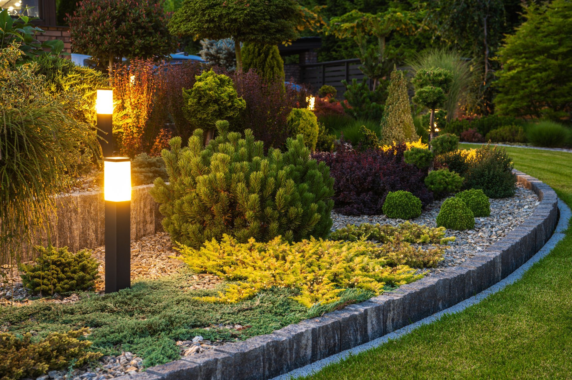 A landscaped garden bed with evergreen shrubs, yellow plants, and lit bollard lights, edged with stone along a lawn.