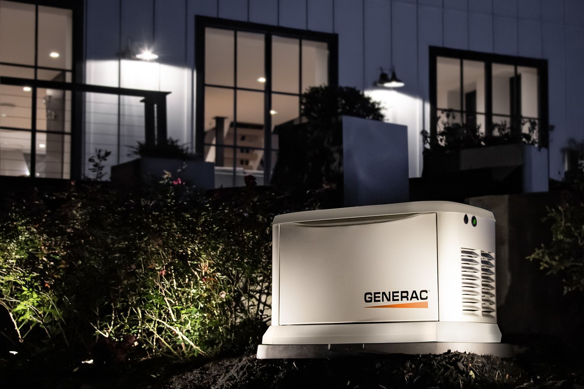 A white Generac standby generator sits in a dark, landscaped yard in front of a house illuminated by outdoor wall lights.