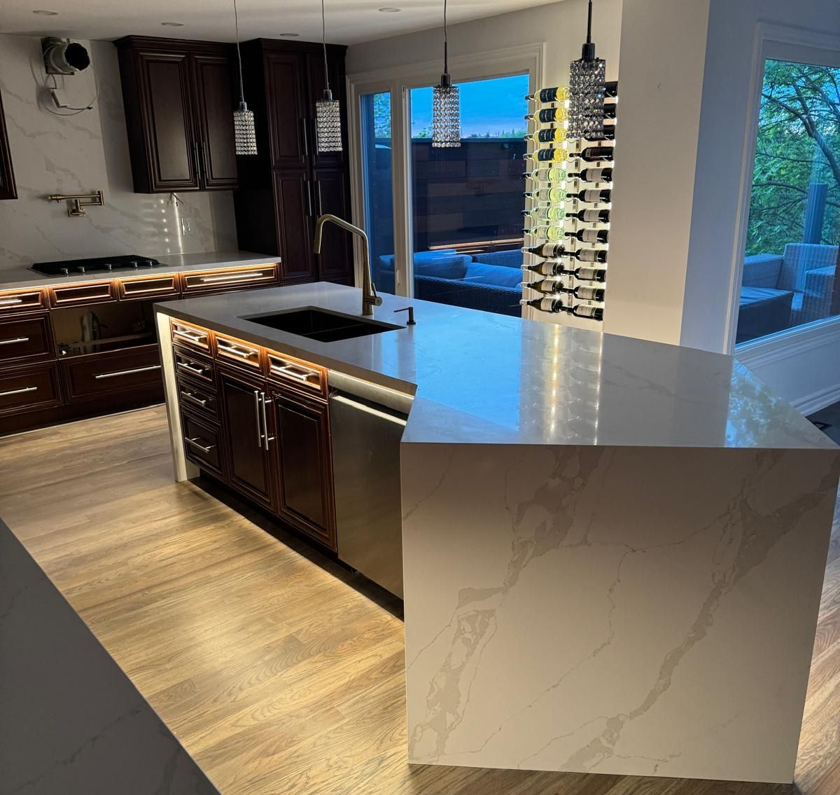 Modern kitchen with island, cabinets, and wine rack, overlooking a patio at dusk.