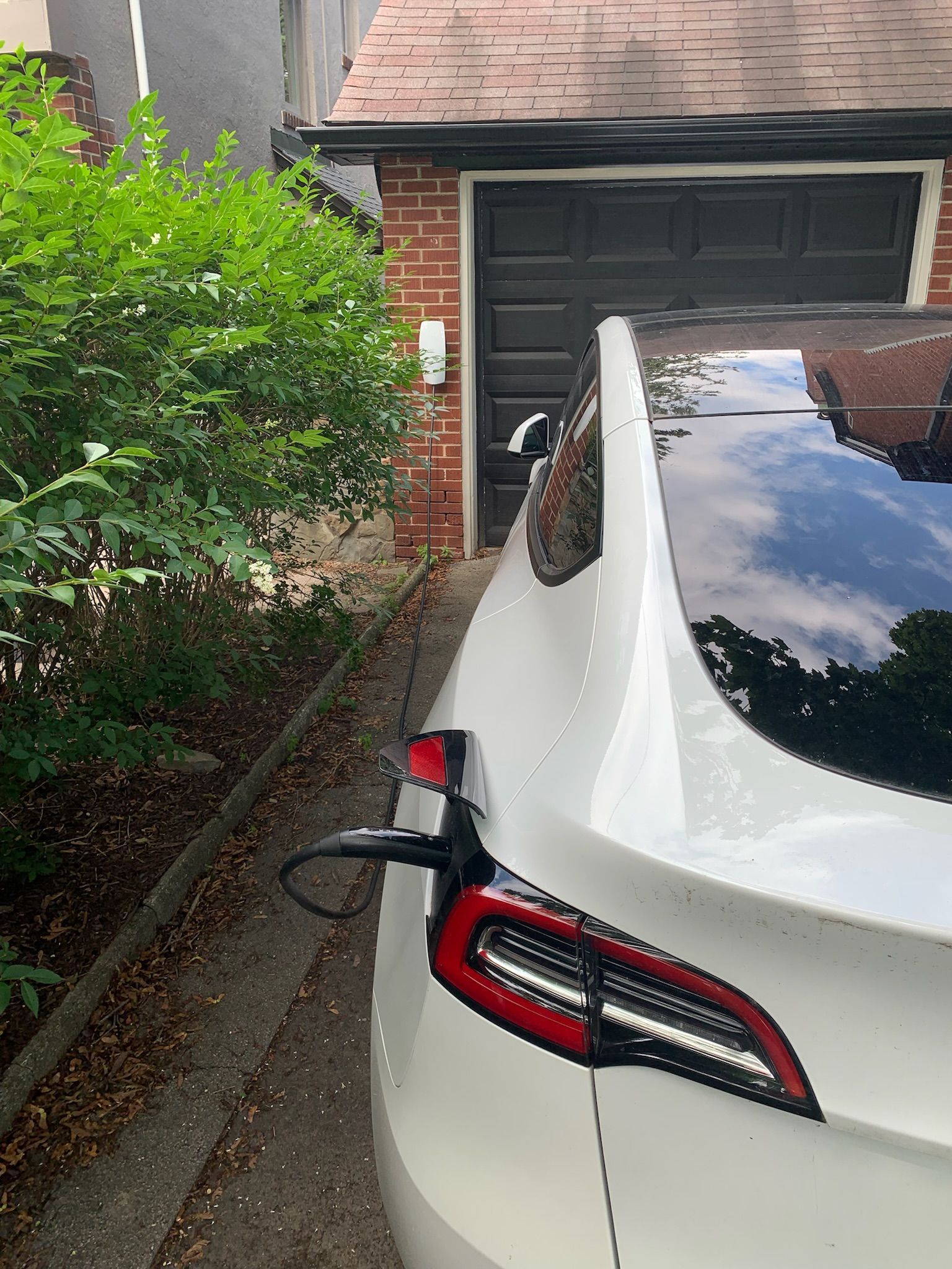 White Tesla charging in a driveway, connected to a wall-mounted charger next to a brick garage.
