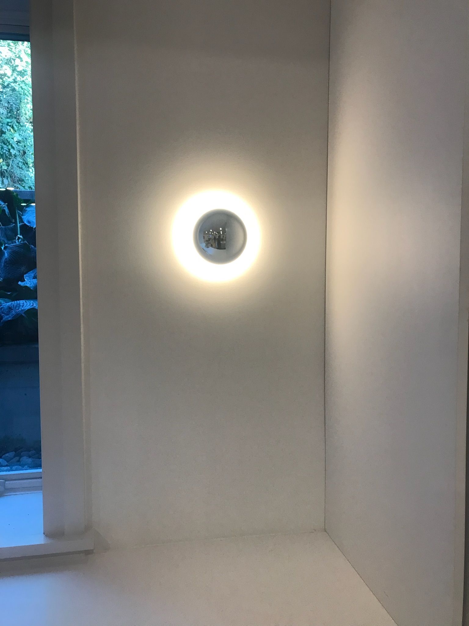 A round, illuminated wall-mounted light fixture on a white wall in a corner near a window.