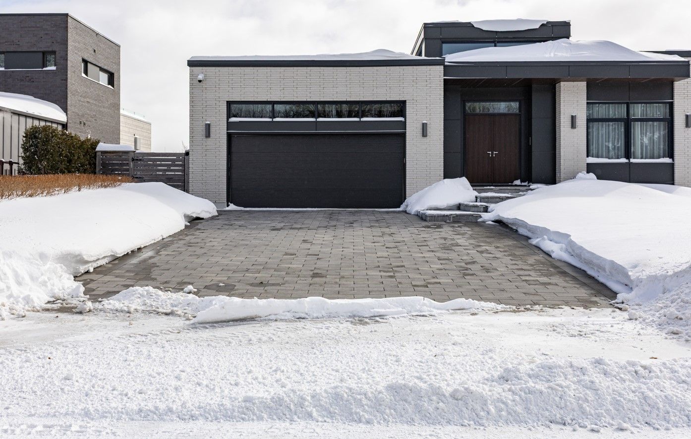 Modern house with snow-covered driveway and garage. The front door is to the right. Snowbanks flank the driveway.