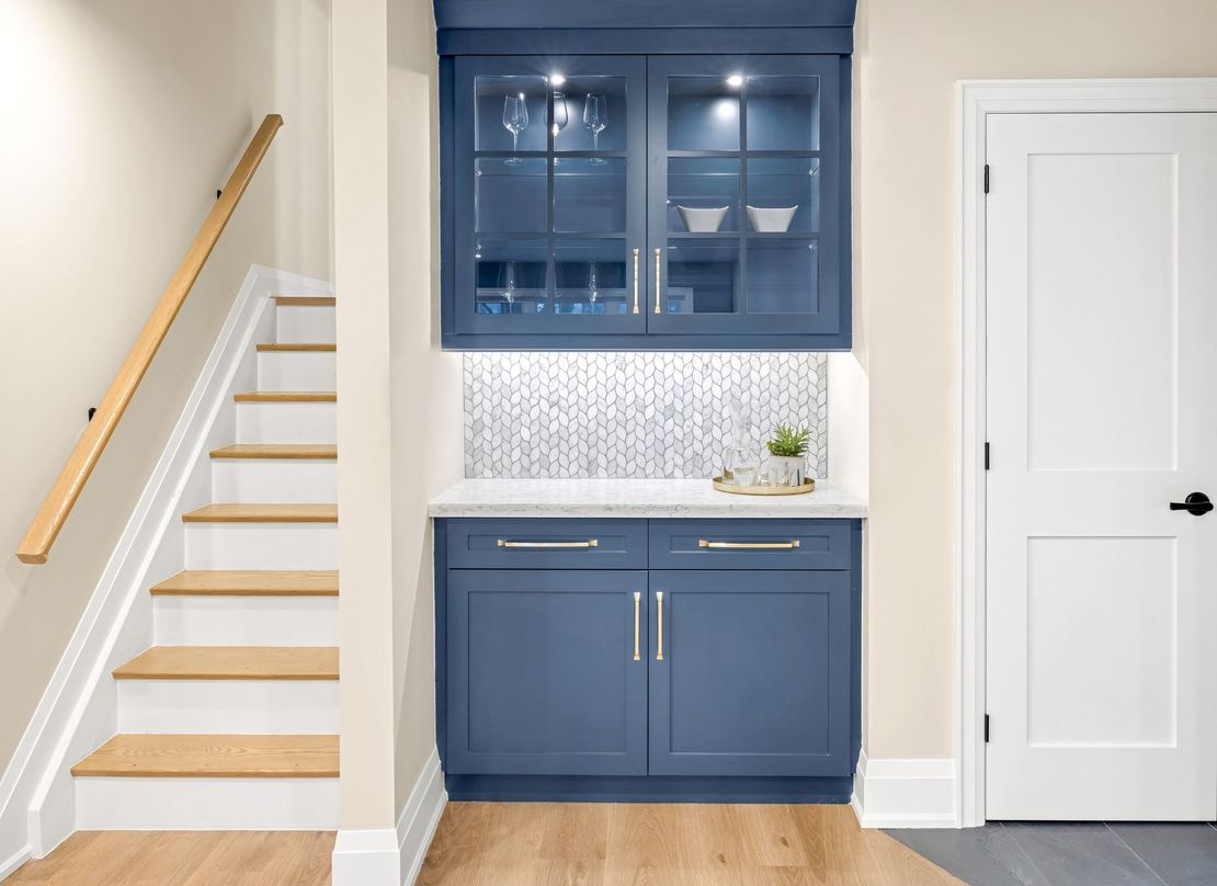 Blue built-in bar with glass cabinets, near a staircase and door in a bright room.
