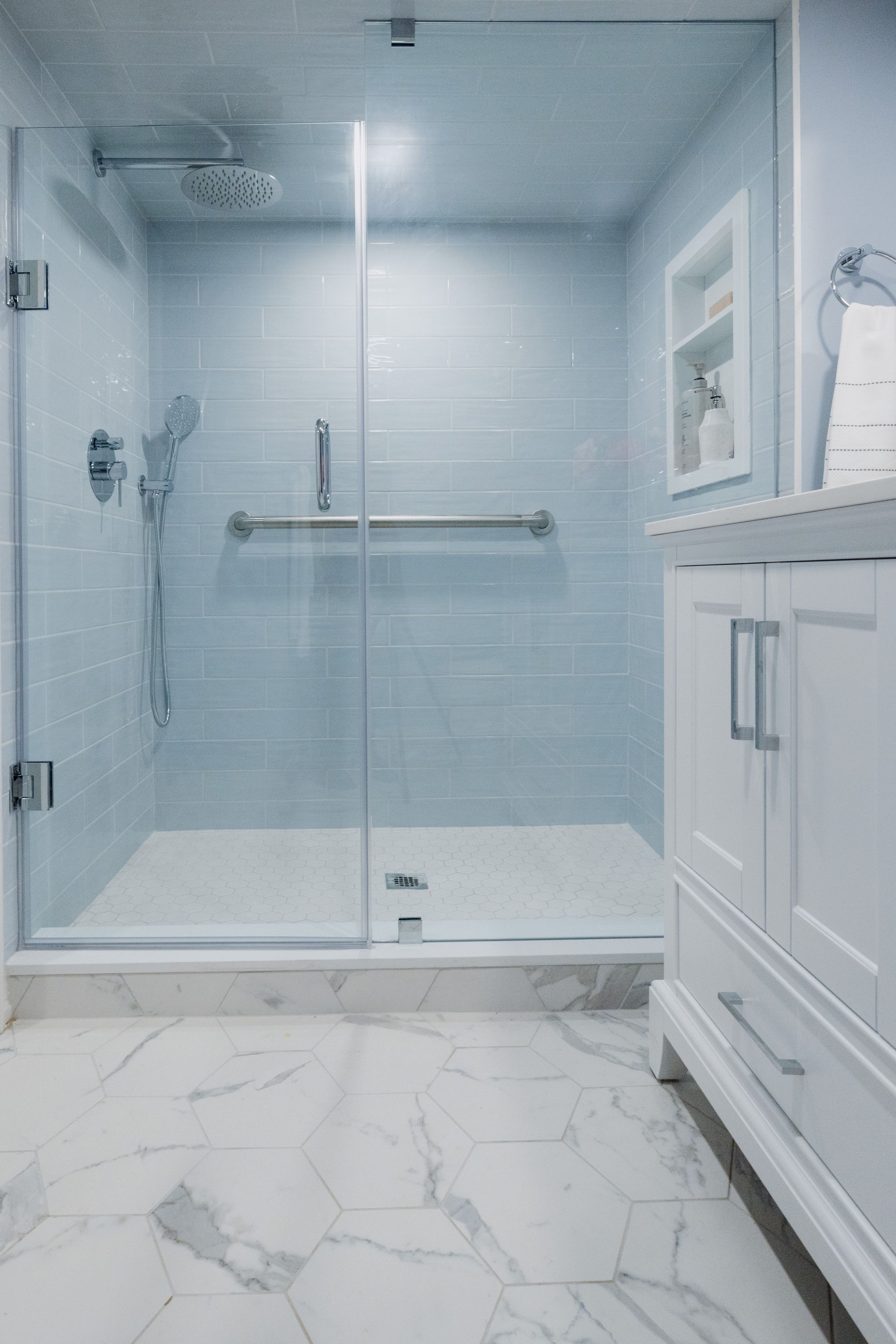 Modern bathroom with glass shower, grab bar, and white vanity on marble floor.