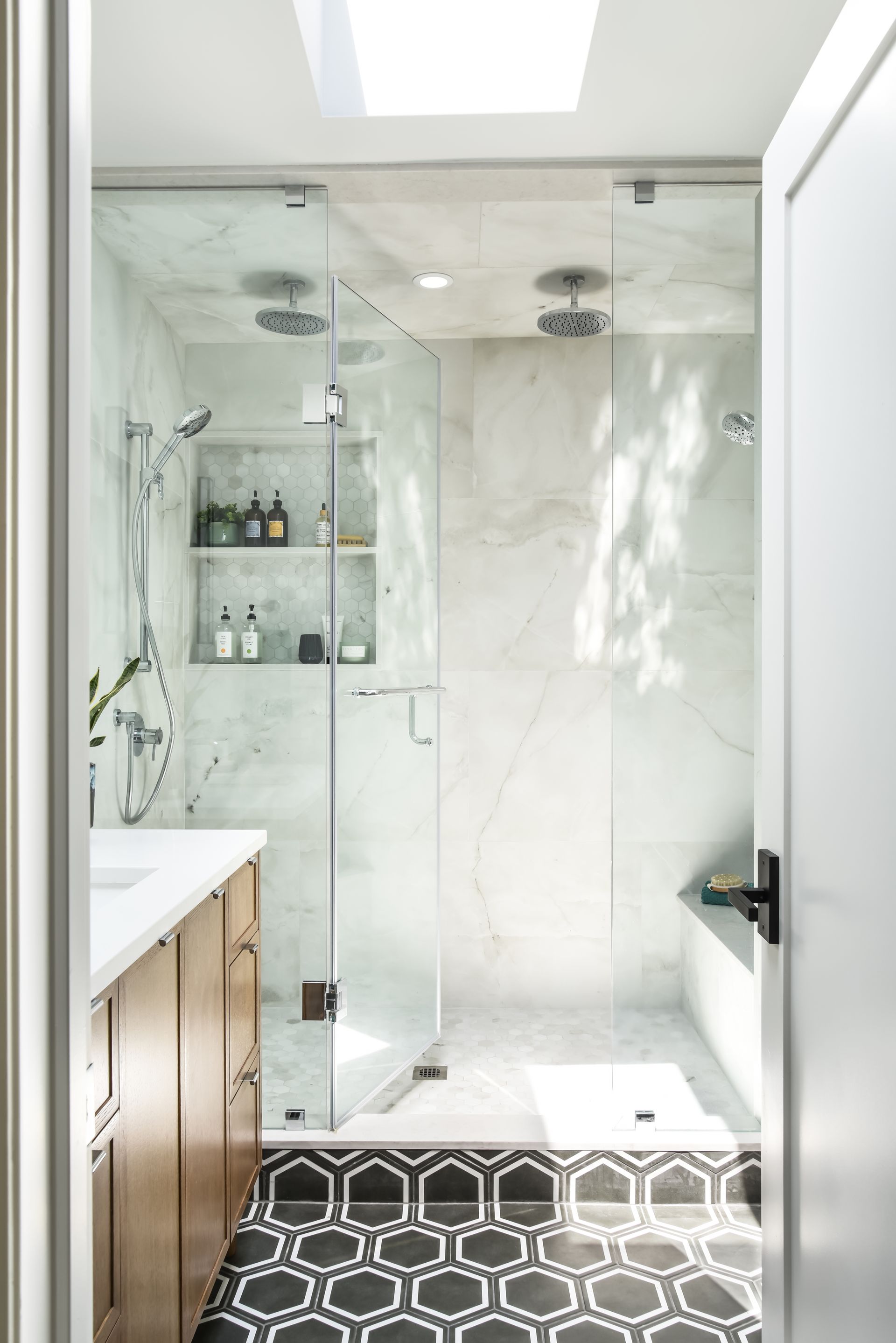 Bathroom with glass shower, patterned floor, and a vanity. Sunlight streams in from a skylight.