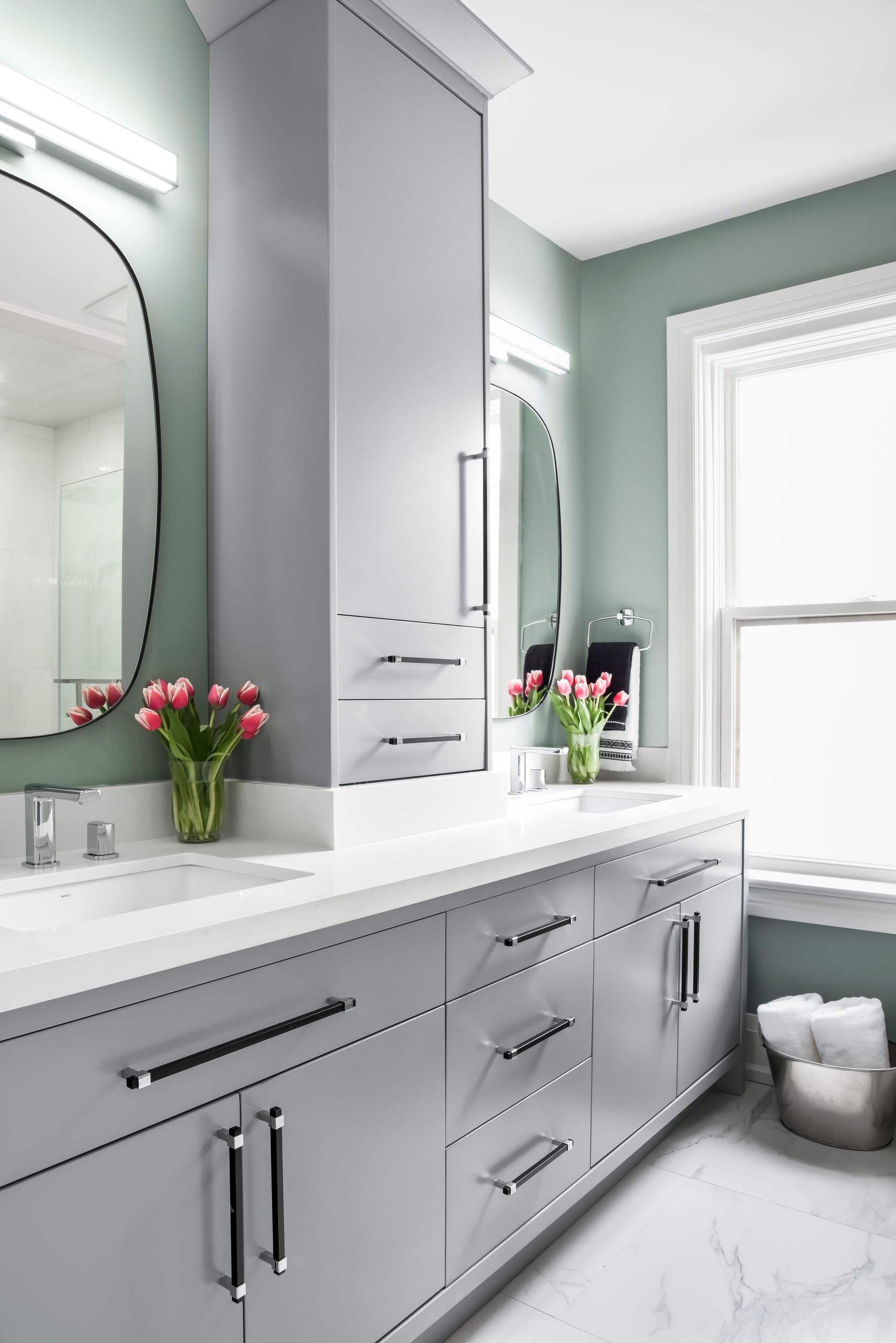 A modern bathroom with a double vanity, a tall cabinet, oval mirrors, and tulips on the countertop.