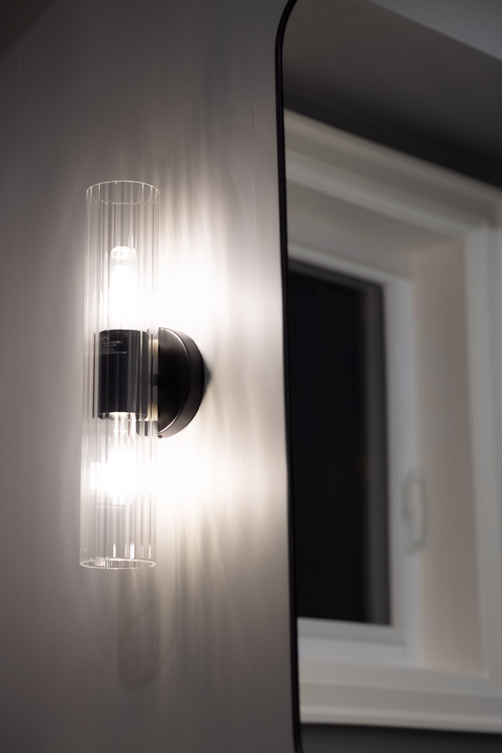 Wall-mounted light fixture with two illuminated glass tubes, next to a window and mirror.