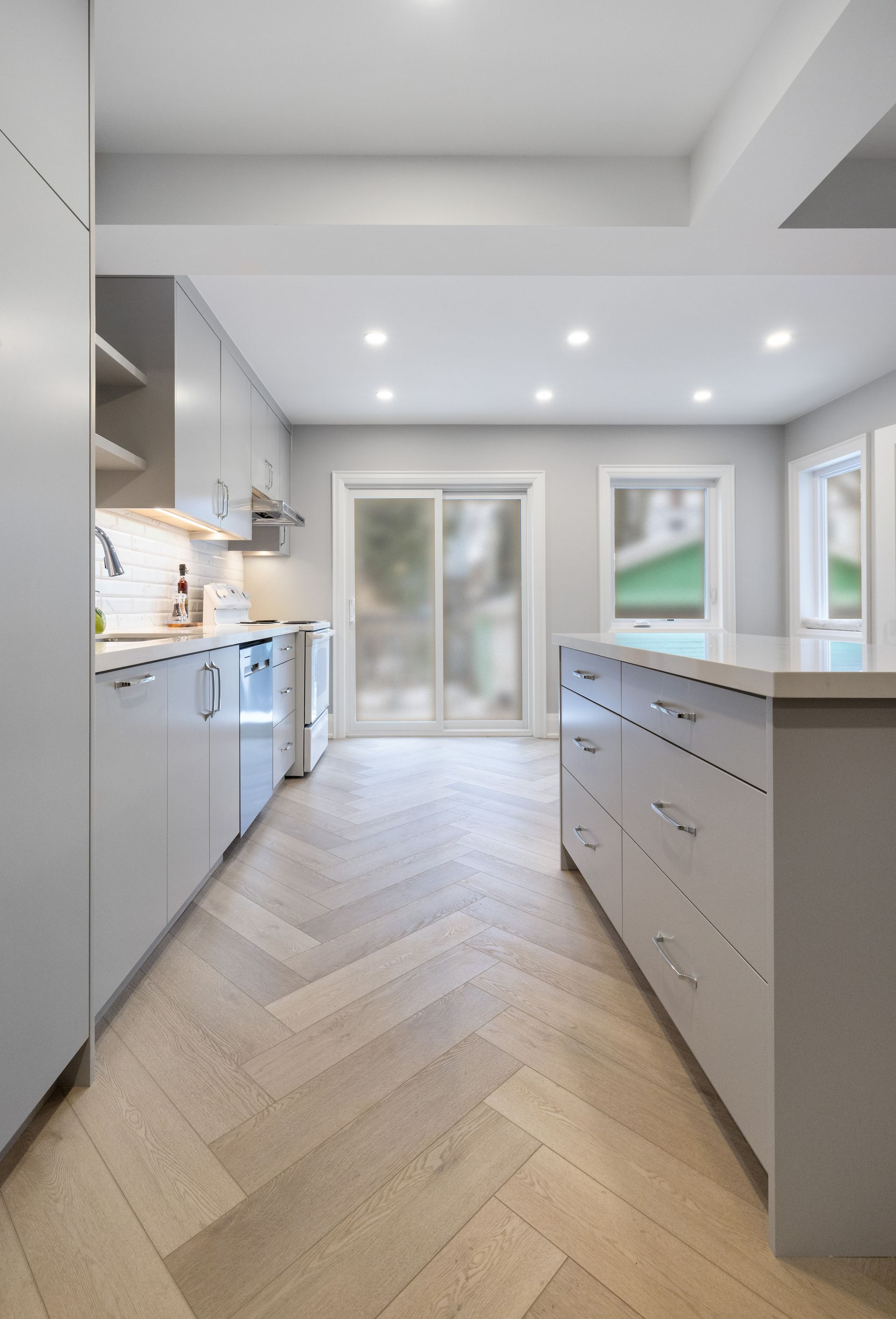 Modern kitchen with light gray cabinets, island, and herringbone wood floor.