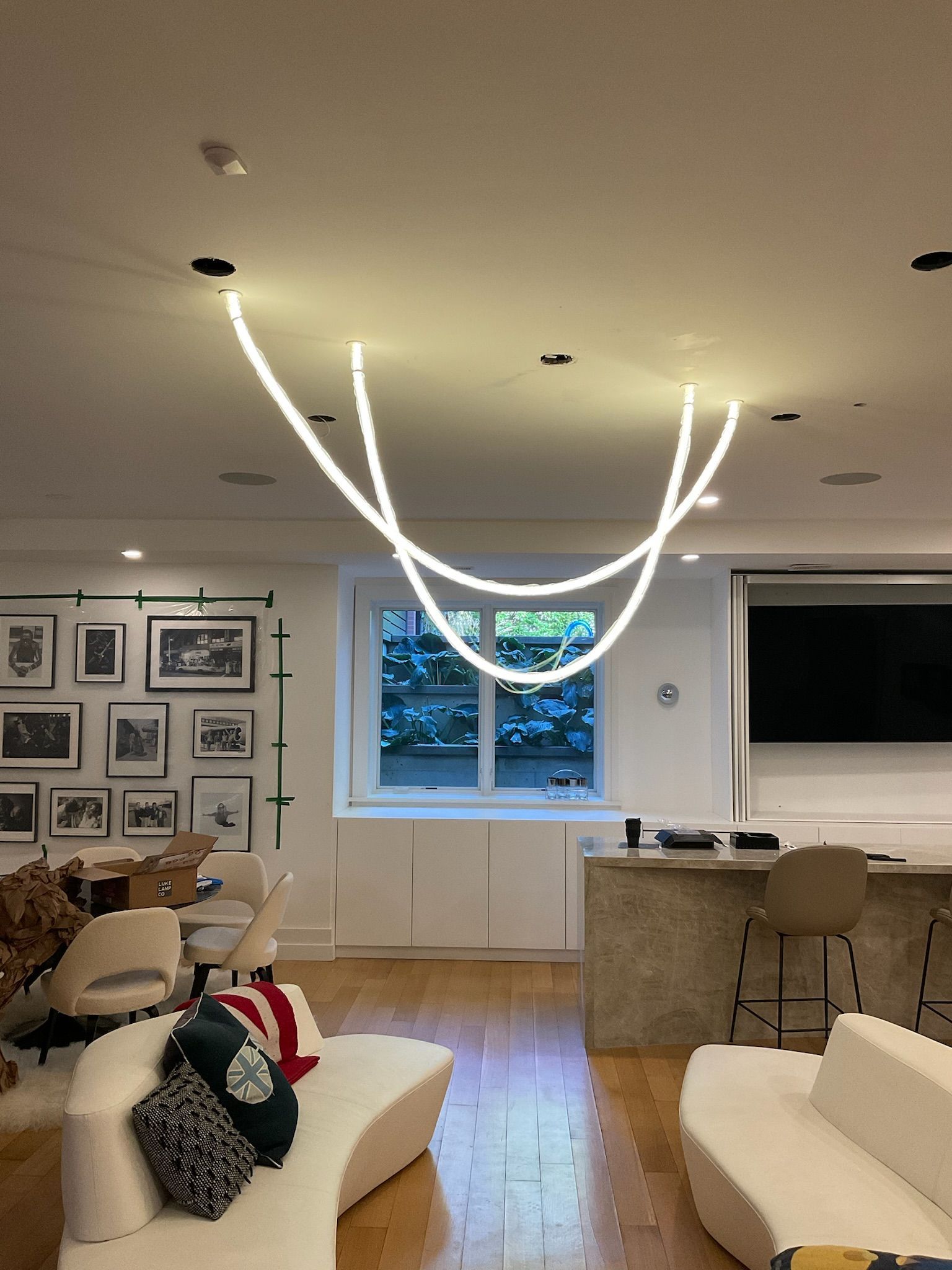 Living room with white sofas, wooden floors, and a curved ceiling light. Window, art, and a bar area are also visible.