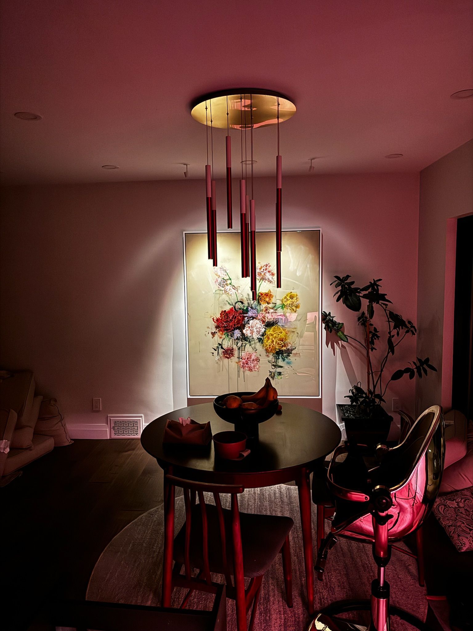 A round table set for a meal beneath a hanging light fixture, with colorful floral art and plants.