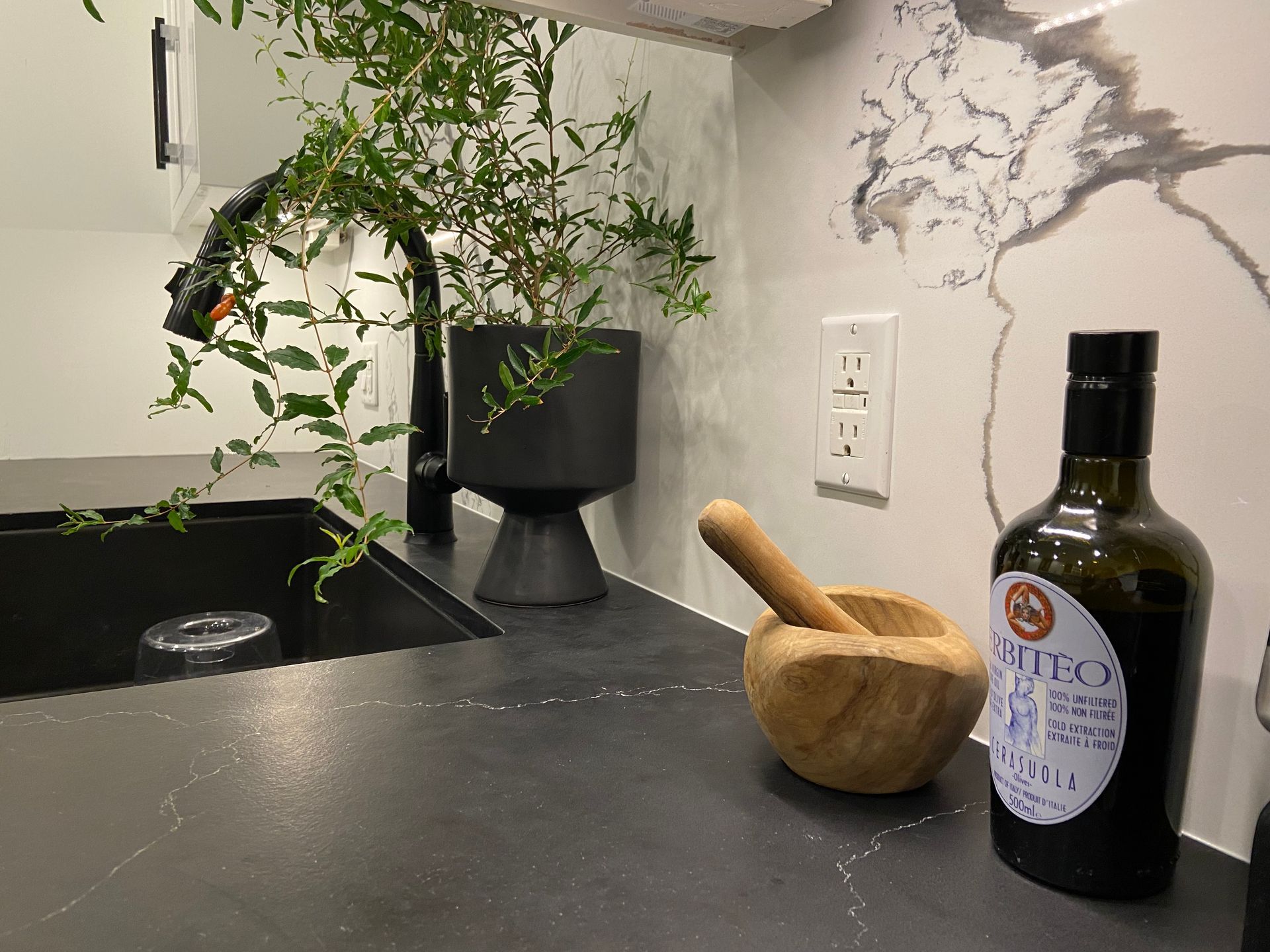 Kitchen countertop with black sink and faucet, plant, mortar and pestle, and olive oil bottle.