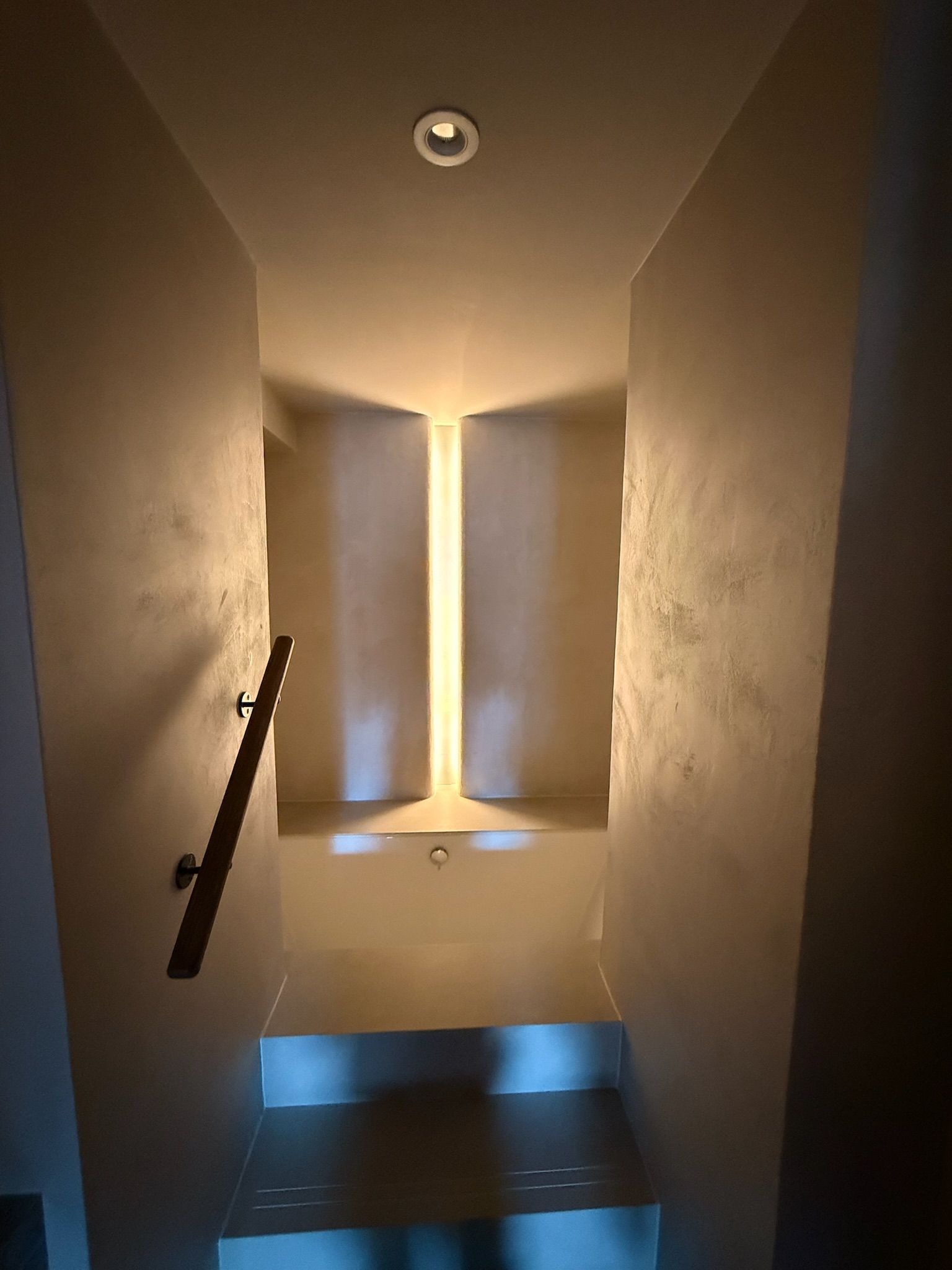 Narrow staircase with a light feature on the wall in the middle. Handrail on the left.