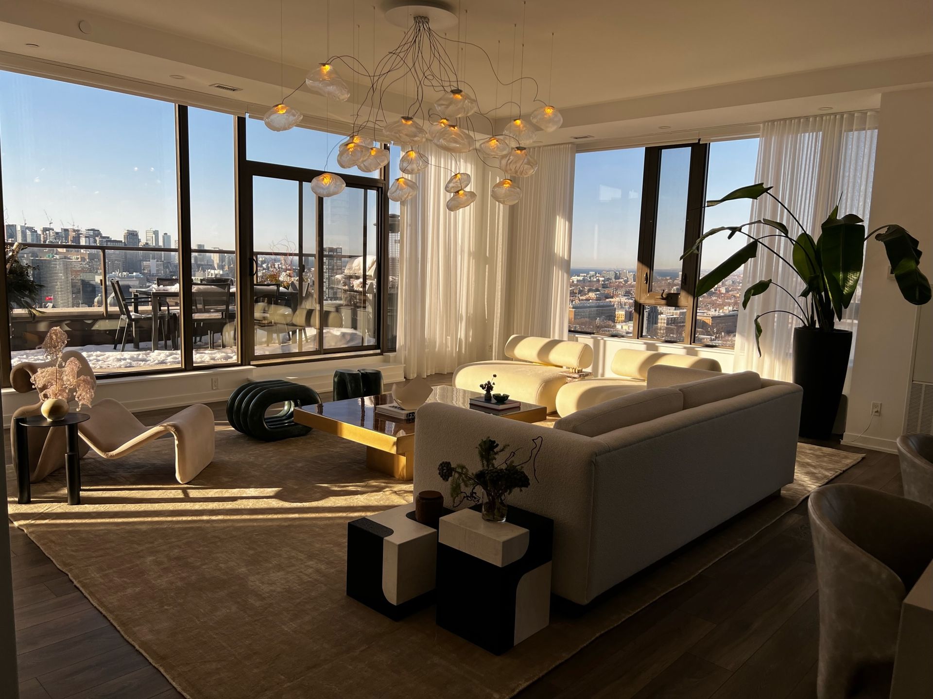 Luxury living room with city view, large windows, white sofa, chandelier, and modern decor.