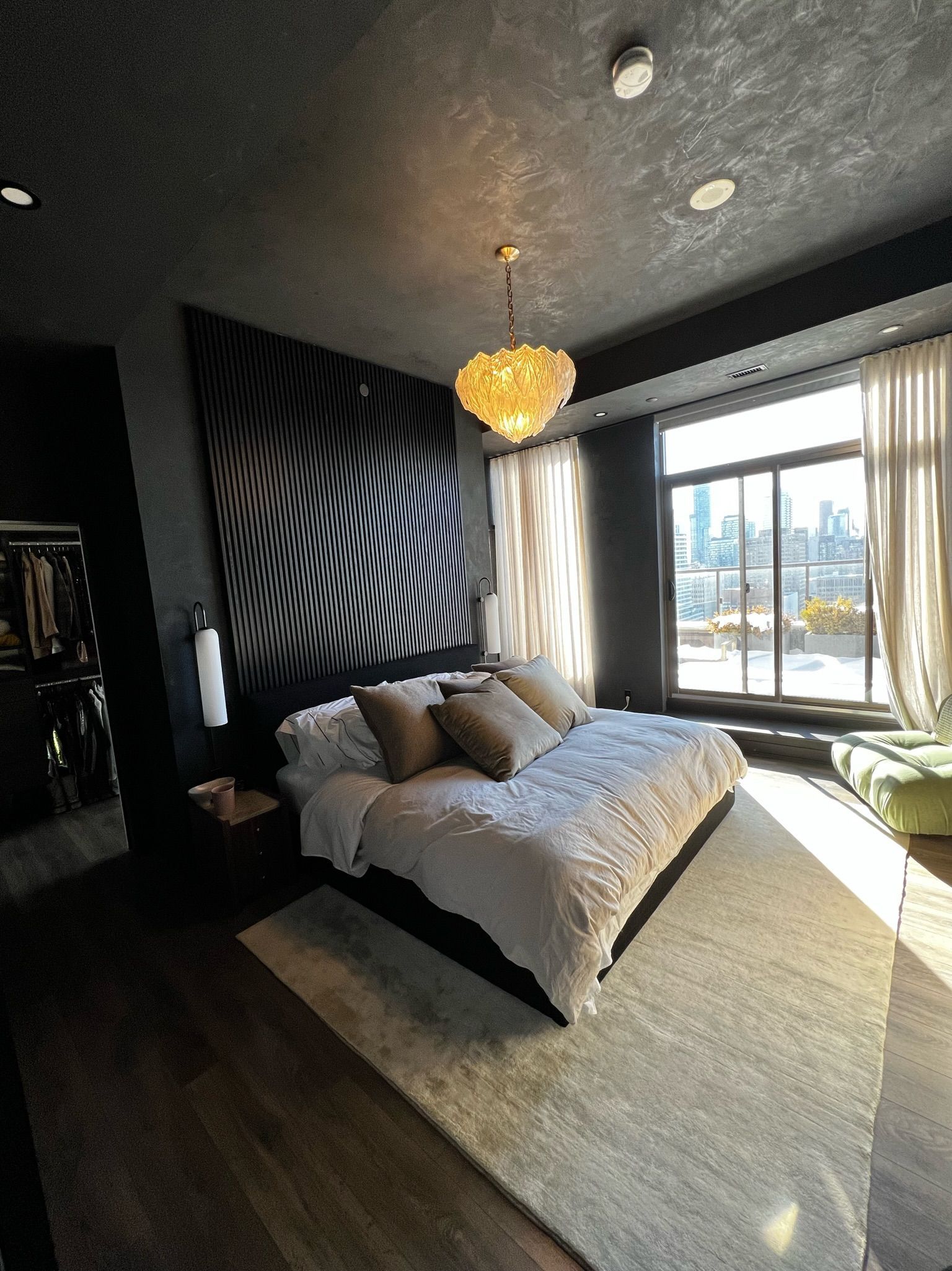Dark bedroom with a bed, patterned headboard, and chandelier. Large window with curtains and a cityscape view.