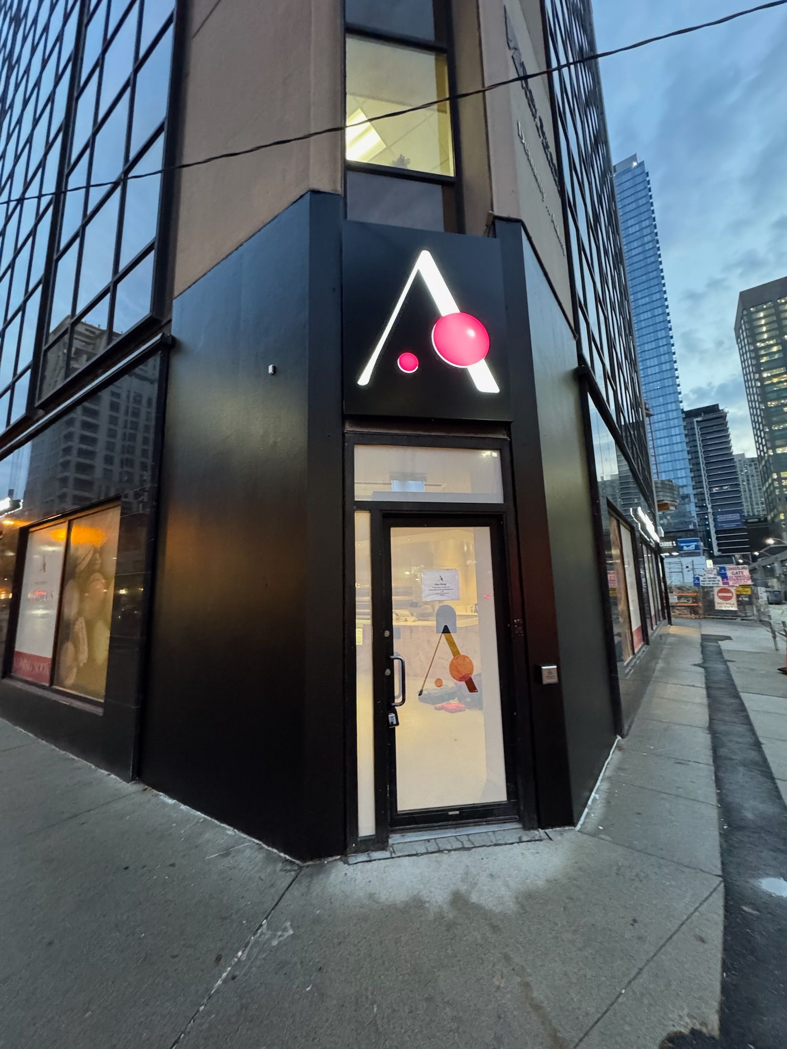 Black-fronted building with a pink and white logo above the entrance; a city street in the background.