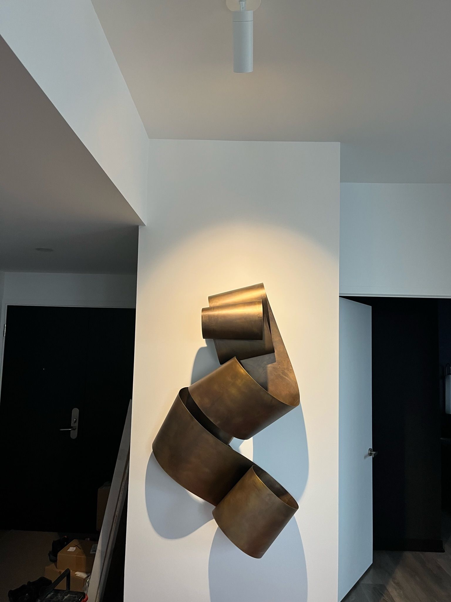Abstract bronze sculpture on white wall, lit by spotlight.