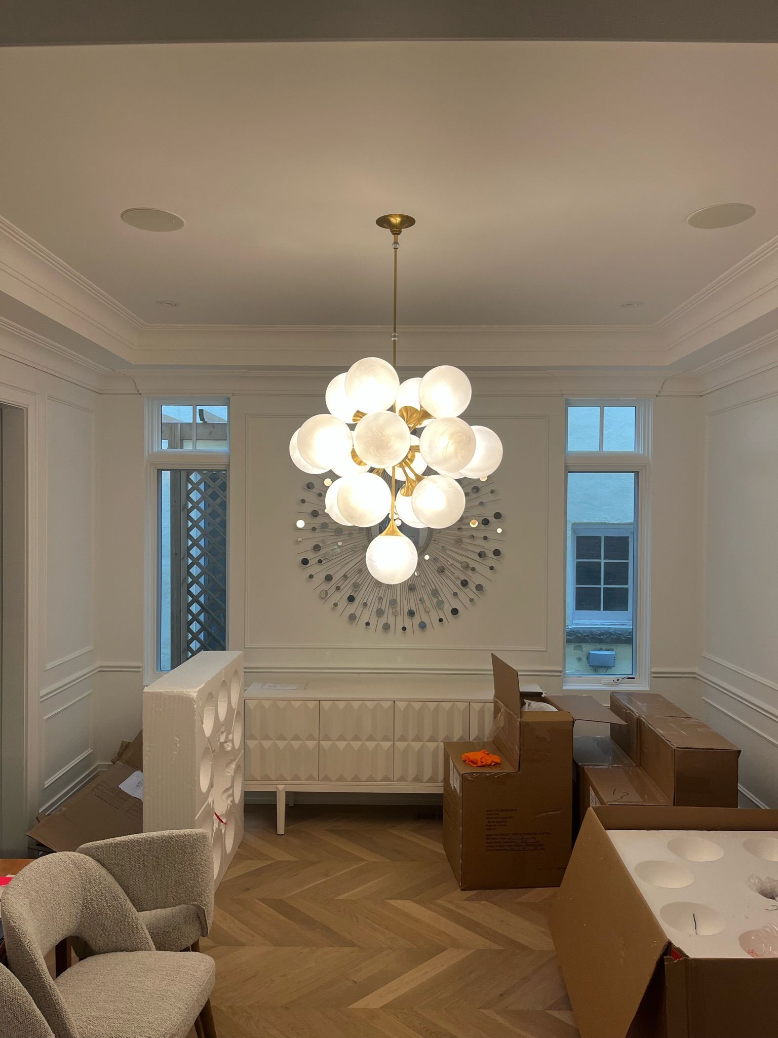Dining room with chandelier, white walls, herringbone wood floor, and boxes.