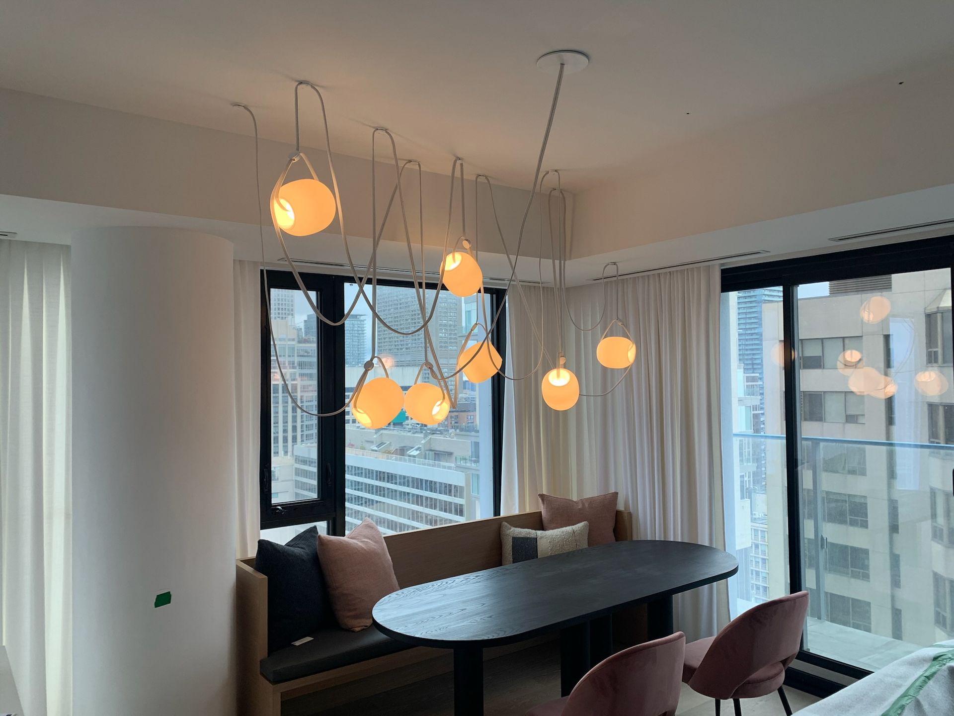 Dining area with black table, built-in bench, pink chairs, large windows, and a multi-bulb pendant light fixture.
