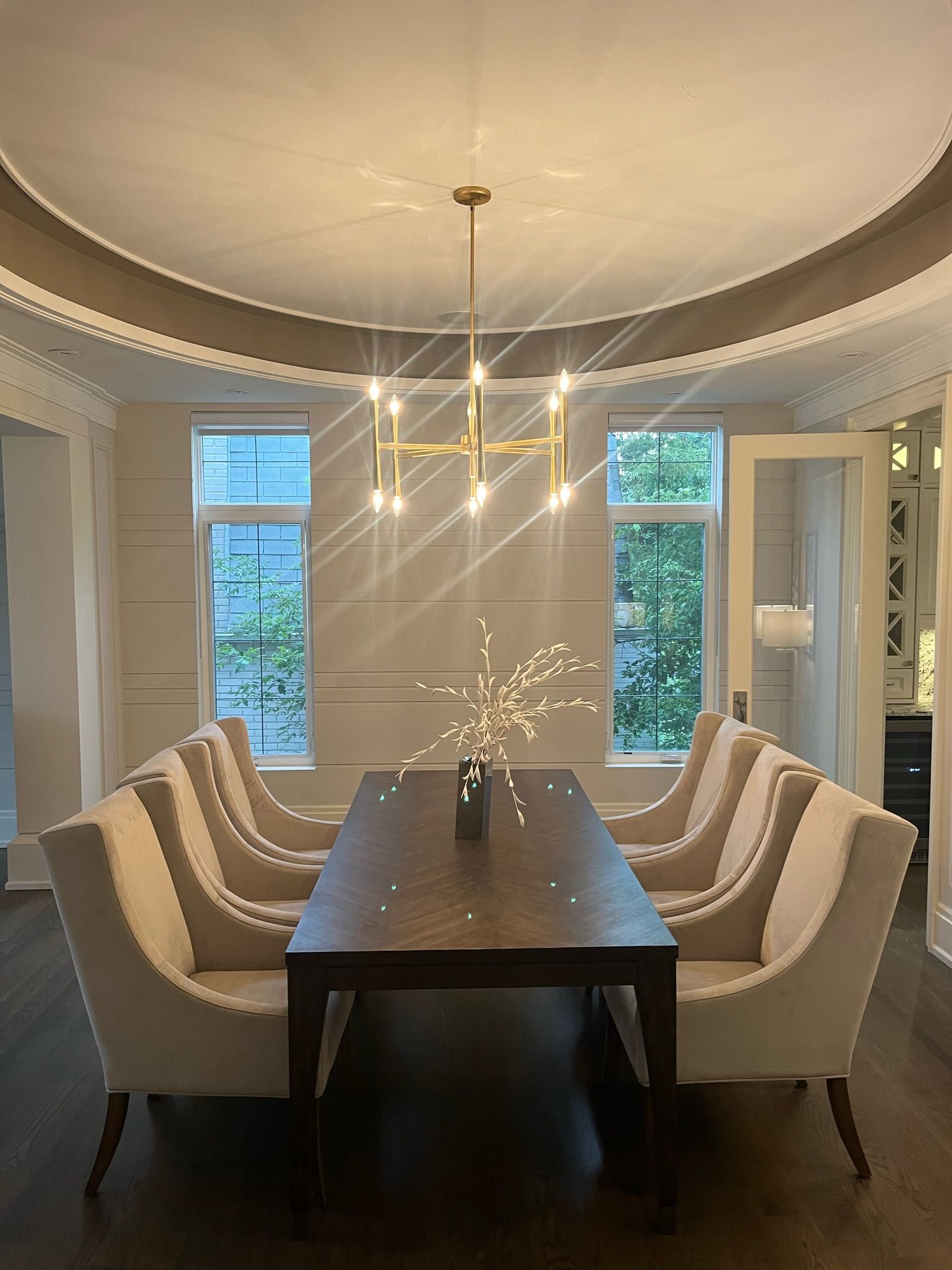 Dining room with a long table, cream chairs, and gold chandelier. Windows and white trim.